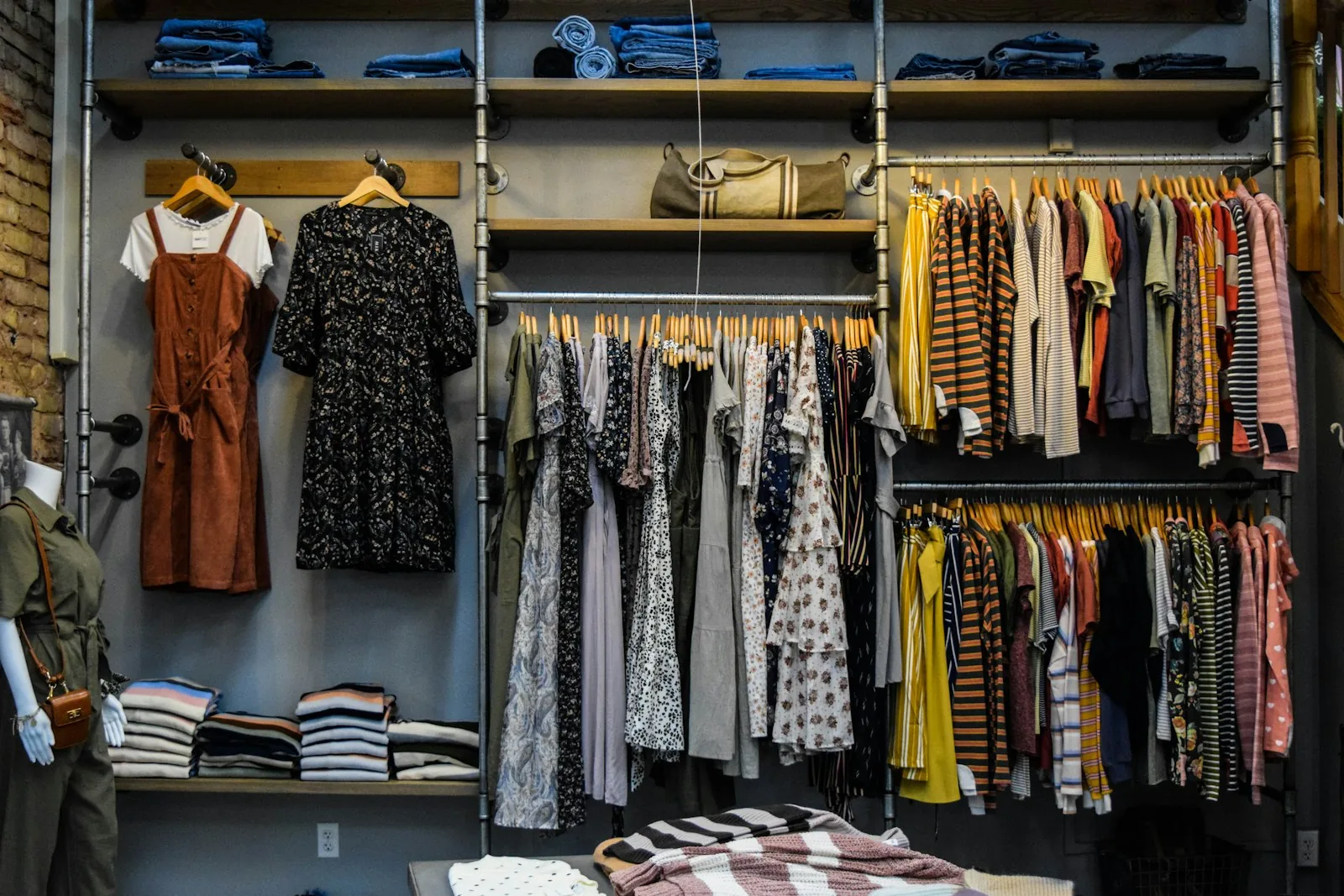 Clothing boutique wall with dresses and striped tops on rails, folded denim on wooden shelves and a mannequin in an olive outfit