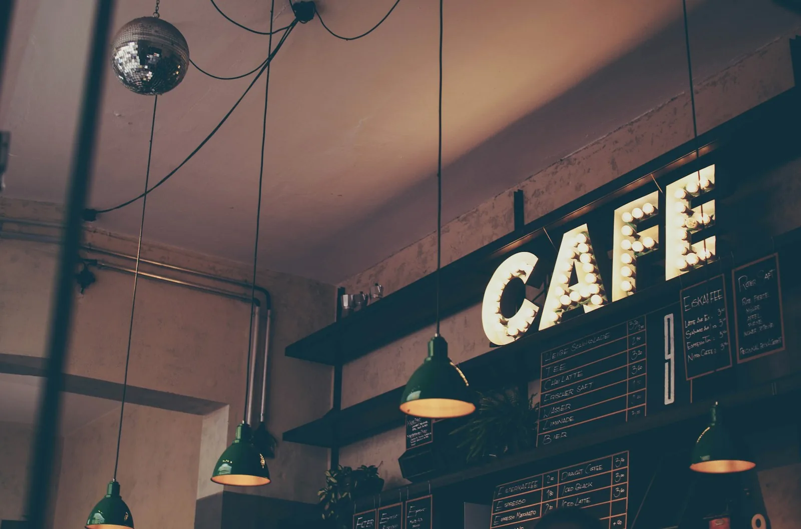 An illuminated CAFE marquee sign above chalkboard drinks menus and green pendant lamps behind a coffee shop counter
